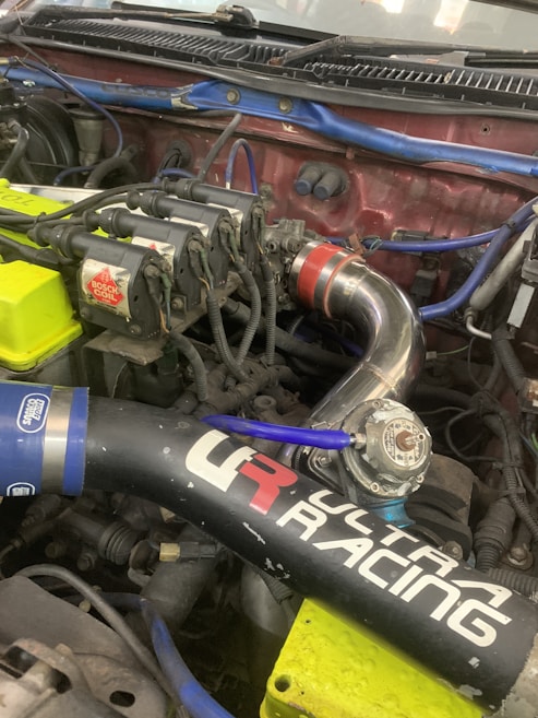 A close-up view of a car engine with visible components including ignition coils marked with 'Bosch Coil', various pipes, and wires. There is a prominent black air intake tube with 'Ultra Racing' branding and colorful wires in blue and yellow. A metallic pipe featuring a red connector is also visible.