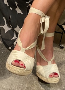A pair of feet wearing white wedge sandals with ankle ties, featuring bright red toenail polish. The sandals have a textured sole, and part of the lower leg is visible, with a black-and-white patterned fabric in the background.