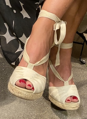 A pair of feet wearing white wedge sandals with ankle ties, featuring bright red toenail polish. The sandals have a textured sole, and part of the lower leg is visible, with a black-and-white patterned fabric in the background.