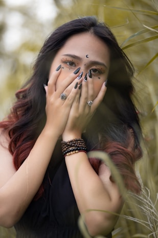 A person with long dark hair is covering their face with their hands, showcasing intricate nail art and facial makeup adorned with stars and crescent shapes. The individual is wearing multiple rings and beaded bracelets, and the background is filled with blurred foliage, giving a natural setting.