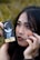 A close-up of a person holding a tarot card labeled 'The Moon' next to their face. The person has celestial symbols like crescent moons and stars drawn around their eyes and forehead. They have black nail polish and long dark hair, and their expression is thoughtful and enigmatic.