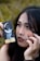 A close-up of a person holding a tarot card labeled 'The Moon' next to their face. The person has celestial symbols like crescent moons and stars drawn around their eyes and forehead. They have black nail polish and long dark hair, and their expression is thoughtful and enigmatic.