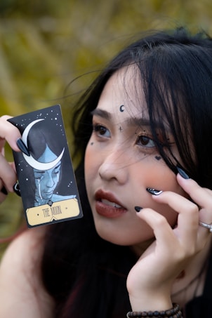 A close-up of a person holding a tarot card labeled 'The Moon' next to their face. The person has celestial symbols like crescent moons and stars drawn around their eyes and forehead. They have black nail polish and long dark hair, and their expression is thoughtful and enigmatic.