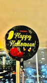 A balloon with the text 'Happy Halloween!' is prominently displayed. The balloon is decorated with Halloween-themed elements including a cute ghost, two pumpkins with faces, candy corn, and spiderwebs. It appears to be indoors, as there are blinds and windows in the background showing an evening cityscape.