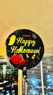 A balloon with the text 'Happy Halloween!' is prominently displayed. The balloon is decorated with Halloween-themed elements including a cute ghost, two pumpkins with faces, candy corn, and spiderwebs. It appears to be indoors, as there are blinds and windows in the background showing an evening cityscape.