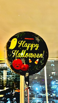A balloon with the text 'Happy Halloween!' is prominently displayed. The balloon is decorated with Halloween-themed elements including a cute ghost, two pumpkins with faces, candy corn, and spiderwebs. It appears to be indoors, as there are blinds and windows in the background showing an evening cityscape.