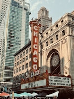A vibrant cityscape features the iconic Chicago Theatre with its bright red and gold marquee sign. Surrounding the theater are tall modern and historic buildings, showcasing a blend of architectural styles. People gather below among colorful umbrellas, suggesting a lively urban atmosphere.