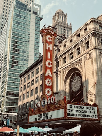 A vibrant cityscape features the iconic Chicago Theatre with its bright red and gold marquee sign. Surrounding the theater are tall modern and historic buildings, showcasing a blend of architectural styles. People gather below among colorful umbrellas, suggesting a lively urban atmosphere.