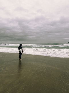 A lone surfer stands on a sandy beach under an overcast sky, gazing at the choppy ocean waves. The surfer is wearing a wetsuit and holding a surfboard, preparing to enter the water. The scene is quiet and introspective, with muted colors reflecting the cloudy weather.