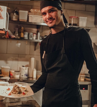 A chef wearing a black apron and black headscarf is standing in a kitchen, holding a white plate with a beautifully plated dish. The background shows various kitchen supplies and equipment on shelves.