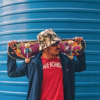 A person wearing a floral bucket hat and sunglasses holds a skateboard across their shoulders. They are dressed in a red t-shirt with 'SKATE BOARD' written on it and a blue jacket. The skateboard features colorful graffiti art. A large blue corrugated metal structure serves as the background.