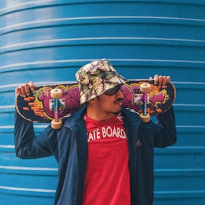 A person wearing a floral bucket hat and sunglasses holds a skateboard across their shoulders. They are dressed in a red t-shirt with 'SKATE BOARD' written on it and a blue jacket. The skateboard features colorful graffiti art. A large blue corrugated metal structure serves as the background.