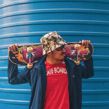 A person wearing a floral bucket hat and sunglasses holds a skateboard across their shoulders. They are dressed in a red t-shirt with 'SKATE BOARD' written on it and a blue jacket. The skateboard features colorful graffiti art. A large blue corrugated metal structure serves as the background.