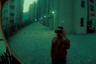 A fisheye lens captures a distorted reflection in a convex mirror, featuring a person taking a photo with a camera. Surrounding the individual are tall buildings in a cityscape, with dim lighting contributing to a moody atmosphere.