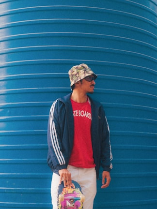A person is standing against a large blue wall, wearing a floral bucket hat, sunglasses, a navy blue jacket with white stripes, a red shirt with 'SKATE BOARD' written on it, and white pants. The person is holding a colorful skateboard with their left hand.