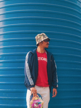 A person is standing against a large blue wall, wearing a floral bucket hat, sunglasses, a navy blue jacket with white stripes, a red shirt with 'SKATE BOARD' written on it, and white pants. The person is holding a colorful skateboard with their left hand.