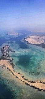 An aerial view of a coastal landscape featuring turquoise waters and sandy islands. The water shows various shades of blue and green, with sandy beaches and small patches of vegetation.