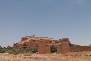 An ancient clay fortress or kasbah set against a clear blue sky, surrounded by sparse vegetation and palm trees. The structure is made of earth-toned bricks and has tall, rustic walls that blend into the rocky landscape. The scene is tranquil and devoid of people.