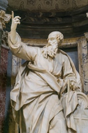A marble statue of a bearded man with an expressive pose, featuring detailed drapery and lifelike facial features. The figure holds an object resembling a book or tablet and stands within an ornately decorated alcove featuring intricate patterns and marble textures.