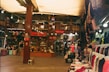A bustling indoor market scene filled with a variety of clothing hanging on racks and mannequins. The shop displays Turkish Delight and other goods. There is a wooden structure with an upper floor, and people are casually browsing.