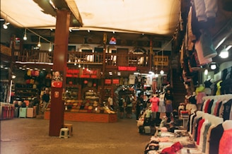 A bustling indoor market scene filled with a variety of clothing hanging on racks and mannequins. The shop displays Turkish Delight and other goods. There is a wooden structure with an upper floor, and people are casually browsing.