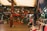 A bustling indoor market scene filled with a variety of clothing hanging on racks and mannequins. The shop displays Turkish Delight and other goods. There is a wooden structure with an upper floor, and people are casually browsing.
