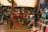 A bustling indoor market scene filled with a variety of clothing hanging on racks and mannequins. The shop displays Turkish Delight and other goods. There is a wooden structure with an upper floor, and people are casually browsing.