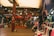 A bustling indoor market scene filled with a variety of clothing hanging on racks and mannequins. The shop displays Turkish Delight and other goods. There is a wooden structure with an upper floor, and people are casually browsing.