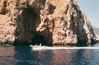 A motorboat glides across clear blue water in front of a large rocky cliff with a cave opening. The cliff has a rugged texture, and patches of vegetation can be seen in certain areas. The boat is occupied by people enjoying the scenery.