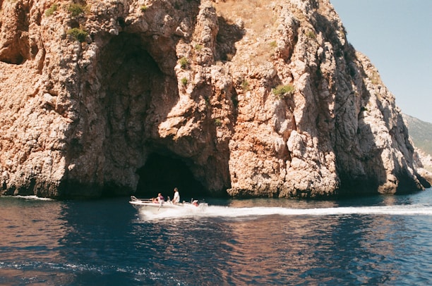 A motorboat glides across clear blue water in front of a large rocky cliff with a cave opening. The cliff has a rugged texture, and patches of vegetation can be seen in certain areas. The boat is occupied by people enjoying the scenery.