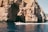 A motorboat glides across clear blue water in front of a large rocky cliff with a cave opening. The cliff has a rugged texture, and patches of vegetation can be seen in certain areas. The boat is occupied by people enjoying the scenery.