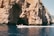 A motorboat glides across clear blue water in front of a large rocky cliff with a cave opening. The cliff has a rugged texture, and patches of vegetation can be seen in certain areas. The boat is occupied by people enjoying the scenery.