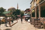 A vibrant street scene featuring a mixture of historical and modern elements. There are several pedestrians walking along a cobblestone street lined with restaurants displaying menus and shops with signs in vibrant colors. Yellow taxis are parked on the left, while a large domed mosque with a tall minaret is visible in the background amidst lush green trees.