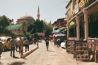 A vibrant street scene featuring a mixture of historical and modern elements. There are several pedestrians walking along a cobblestone street lined with restaurants displaying menus and shops with signs in vibrant colors. Yellow taxis are parked on the left, while a large domed mosque with a tall minaret is visible in the background amidst lush green trees.
