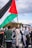 A crowd of people participating in a protest, with one individual prominently holding a large flag featuring black, white, green, and red sections. Several other participants are visible, some holding signs with messages such as 'End the blockade on Gaza'. The scene takes place outdoors with partly cloudy skies visible above.