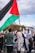 A crowd of people participating in a protest, with one individual prominently holding a large flag featuring black, white, green, and red sections. Several other participants are visible, some holding signs with messages such as 'End the blockade on Gaza'. The scene takes place outdoors with partly cloudy skies visible above.