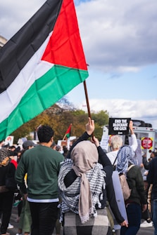 A crowd of people participating in a protest, with one individual prominently holding a large flag featuring black, white, green, and red sections. Several other participants are visible, some holding signs with messages such as 'End the blockade on Gaza'. The scene takes place outdoors with partly cloudy skies visible above.