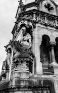 An ornate stone mausoleum featuring intricate architectural details and two statues. The foreground statue depicts a seated figure in a draped garment with a solemn expression. The stone structure includes decorative carvings and gothic elements, creating a historical and somber atmosphere.