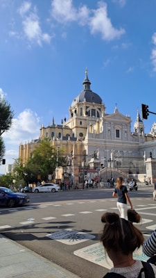 A large, ornate cathedral with multiple domes and spires is set against a bright blue sky with scattered clouds. People are walking near the building, and cars are present on the street. Trees and traffic signals add to the urban setting in front of the cathedral.