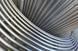 Interlocking stainless steel tubes form an abstract pattern, with a smooth metallic texture. The arrangement creates curved, overlapping lines that suggest movement and flow. The background is a neutral, concrete surface.