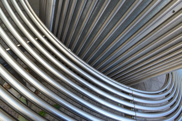Interlocking stainless steel tubes form an abstract pattern, with a smooth metallic texture. The arrangement creates curved, overlapping lines that suggest movement and flow. The background is a neutral, concrete surface.