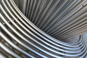 Interlocking stainless steel tubes form an abstract pattern, with a smooth metallic texture. The arrangement creates curved, overlapping lines that suggest movement and flow. The background is a neutral, concrete surface.