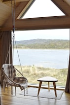 A cozy interior features a hanging chair with a plaid blanket in a wooden cabin. The triangular window provides a view of a serene lake and distant hills. A small, angled wooden table sits beside the chair on a woven rug, complementing the natural tones of the room.