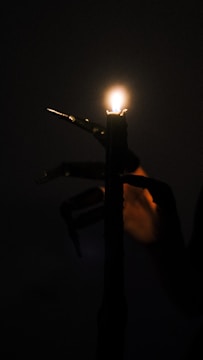 Close-up of trembling hands clutching a flickering candle in pitch darkness.