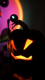 A carved pumpkin with a glowing face is set against a dark background. The pumpkin has sharp, triangular eyes and a jagged mouth, illuminated by an orange light from within. In the background, there is a colorful gradient of light, ranging from purple to green, possibly from another light source.