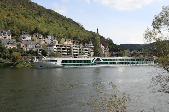 A large, modern river cruise ship sails along a wide river with a backdrop of a quaint village nestled against lush green hills. The village features traditional European architecture with houses and buildings displaying a mix of pastel and neutral colors. Trees and foliage surround the area, adding to the picturesque nature of the scene.