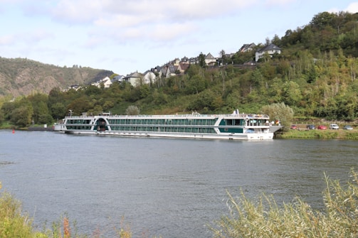 A large river cruise ship is sailing on a wide river bordered by lush green vegetation. On the opposite bank, a hillside is dotted with buildings, houses, and cars. The sky is partly cloudy with patches of blue showing through.