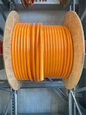 A large wooden spool holds tightly wound coils of thick, vibrant orange cable. The spool is situated on a metal rack in what appears to be an industrial or storage setting. The sheen on the cable suggests it is made of a durable material, possibly for electrical or communication purposes.