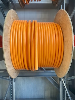 A large wooden spool holds tightly wound coils of thick, vibrant orange cable. The spool is situated on a metal rack in what appears to be an industrial or storage setting. The sheen on the cable suggests it is made of a durable material, possibly for electrical or communication purposes.