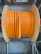 A large wooden spool holds tightly wound coils of thick, vibrant orange cable. The spool is situated on a metal rack in what appears to be an industrial or storage setting. The sheen on the cable suggests it is made of a durable material, possibly for electrical or communication purposes.